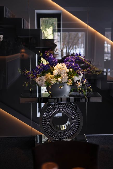 A floral arrangement featuring white roses and purple flowers, possibly delphiniums or hydrangeas, in a smooth, round, white ceramic vase. The bouquet is lush and cascading with greenery, displayed on