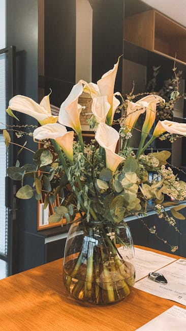 A floral arrangement featuring white roses and purple flowers, possibly delphiniums or hydrangeas, in a smooth, round, white ceramic vase. The bouquet is lush and cascading with greenery, displayed on