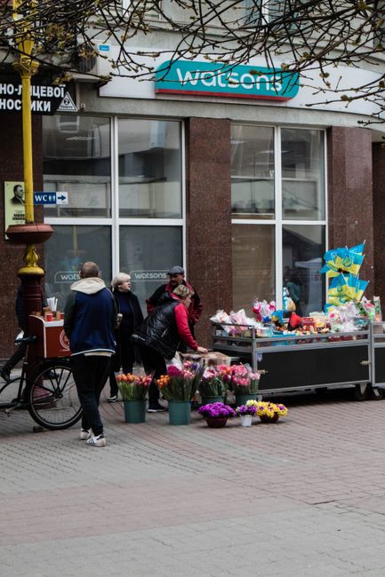 A charming flower display outside a florist shop, featuring a wooden cart filled with vibrant bouquets wrapped in pink and white paper. Surrounding the cart are potted red, pink, and white flowering p